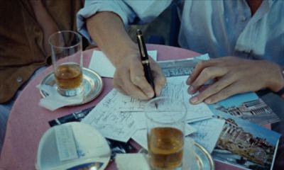 This may contain: two people sitting at a table with papers and glasses on top of it, writing