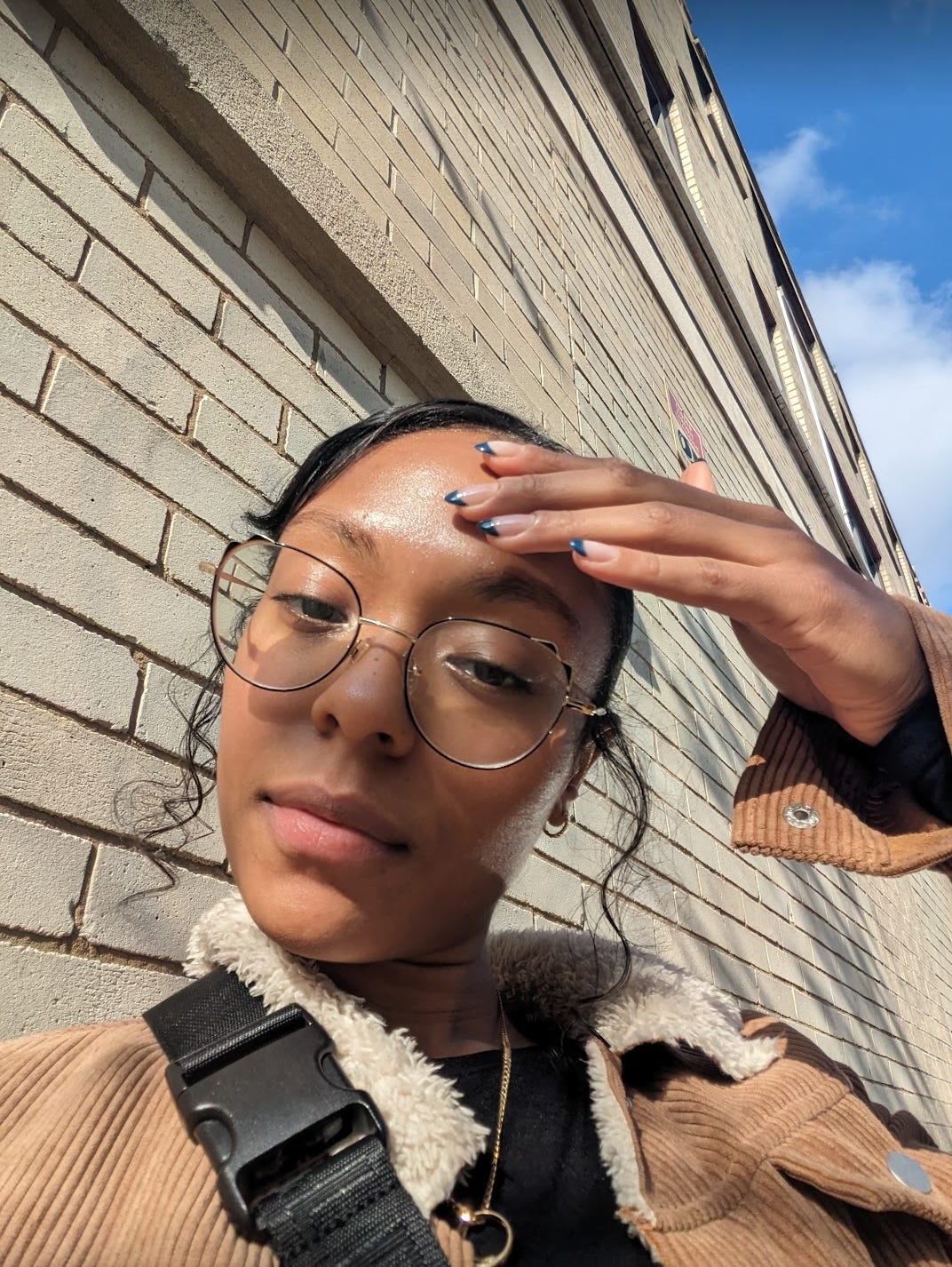  Nathalie takes a selfie showing her glowing brown skin during a very sunny day. She places her hand on her forehead to show off her navy blue french tip gel nails. She has curly strands of her sticking out on either side of her ears to frame her face. She has cat shaped rim glasses, a tan corduroy jacket and a gold necklace peeking out.