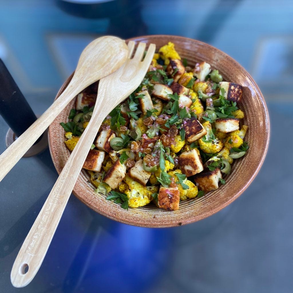 A stoneware serving bowl full of the salad described above: turmeric-tinted roasted cauliflower, browned cubes of halloumi, raisins, shallots, and parsley scattered across the top. Wooden serving utensils rest at the edge of the bowl.