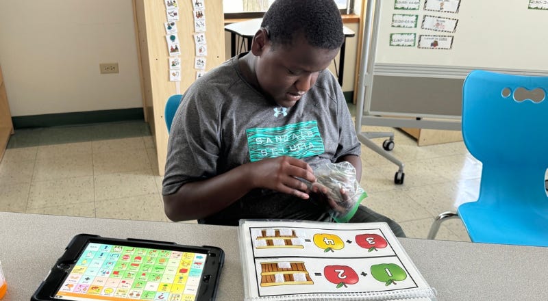 Student looking at a plastic bag. Worksheet and AAC device are on a desk in front of him