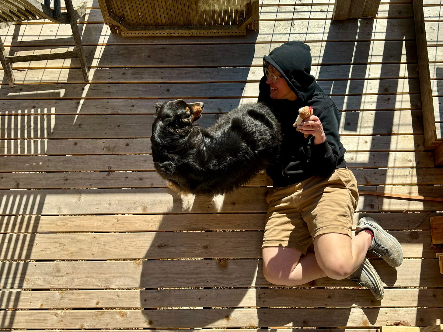 Lewis (extremely adorable black dog) and Beck on a wooden deck. Beck is holding a dog toy.