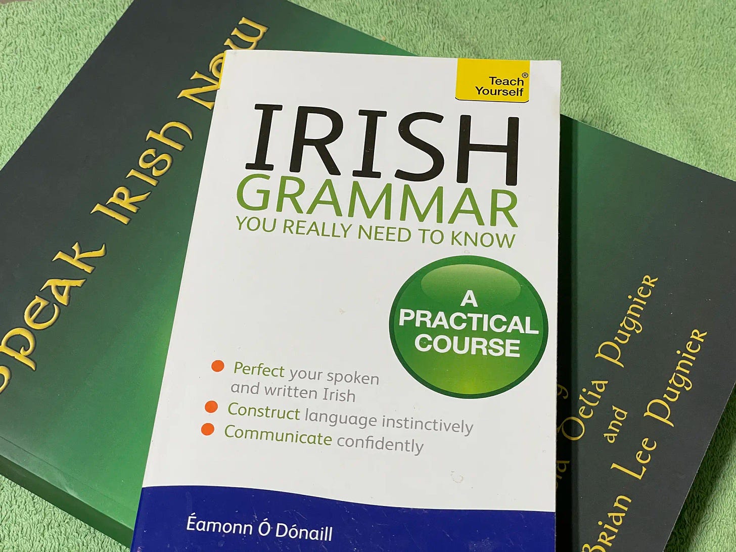 An image of a white book, “Irish Grammar You Really Need to Know”, arranged vertically laid on top of a green book, “Speak Irish Now”.An image of a white book, “Irish Grammar You Really Need to Know”, arranged vertically laid on top of a green book, “Speak Irish Now”.