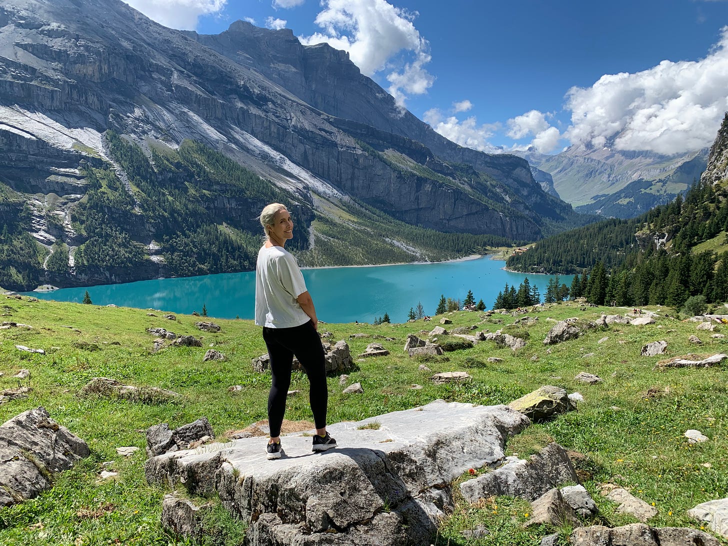 A white woman with blonde hair looks back to smile at the camera with a scenic, bright turquoise lake, mountains, trees, and grass beyond her.