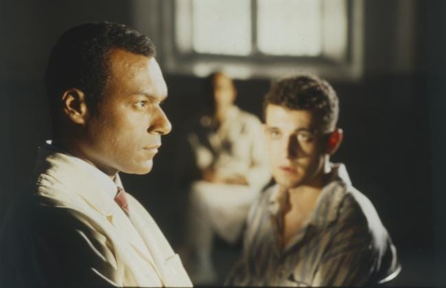 a film still from Isaac Julien's 1995 film Frantz Fanon: Black Skin, White Mask features a Black man in the foreground and a white man in the background and slightly out of focus