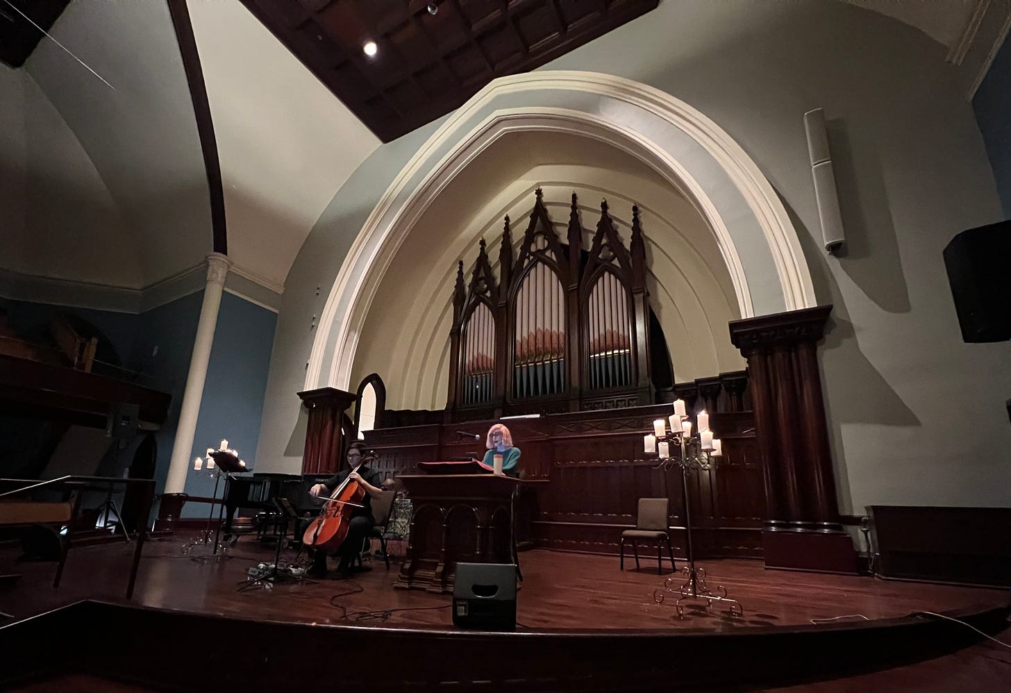 Elle sitting behind pulpit and Julian holding their cello on the "stage" in Sanctuary Hall with the gran organ behind them and candelabras flanking them