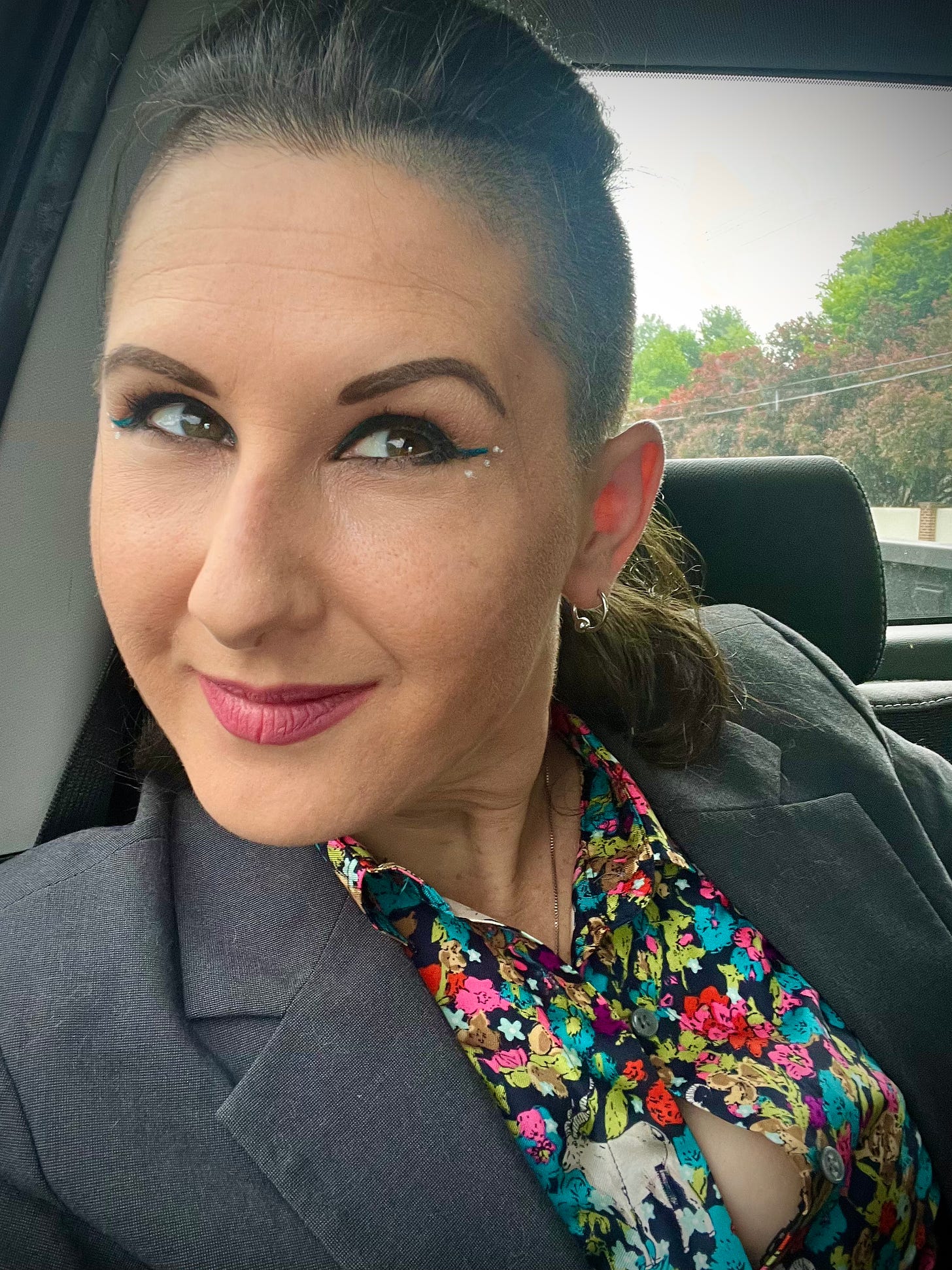 Adult Lyric is in a gray blazer and floral goat shirt. They have winged eyeliner and makeup on as they ride in the back seat of a truck, smiling. 