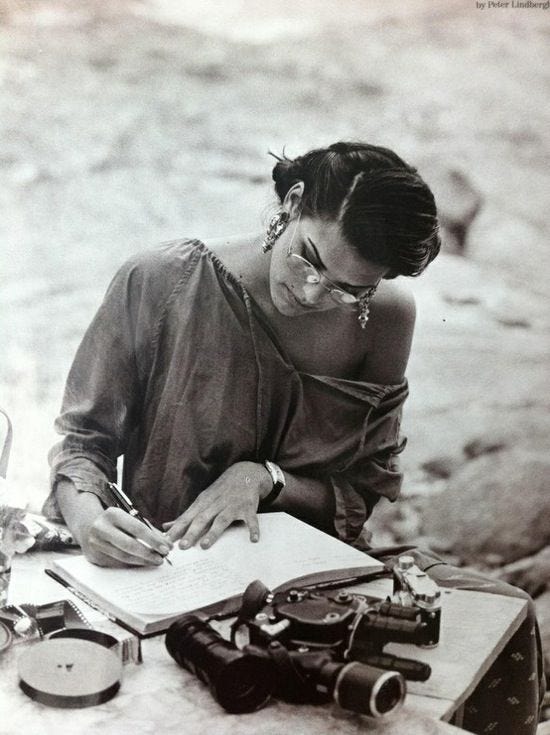 This may contain: a woman sitting at a table writing in a book with old cameras and other items around her