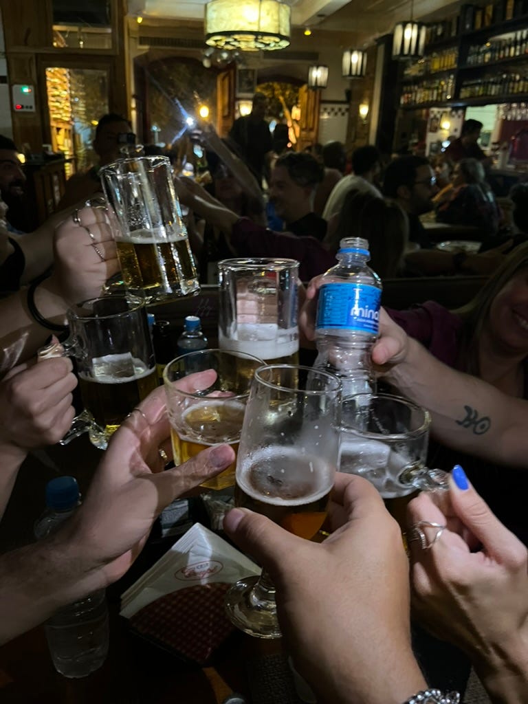 foto em close de sete mãos brindando em bar à noite com copos e taças e bebidas diferentes.