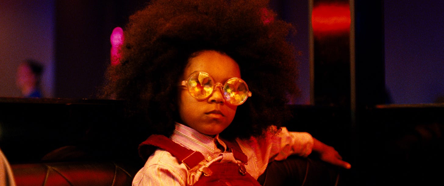A young Black girl sits in a booth, wearing red overalls and a striped button-up shirt, with her arm slung over the back of the booth. Her afro frames her serious face, while wearing prisimed round glasses, as she looks off camera. 