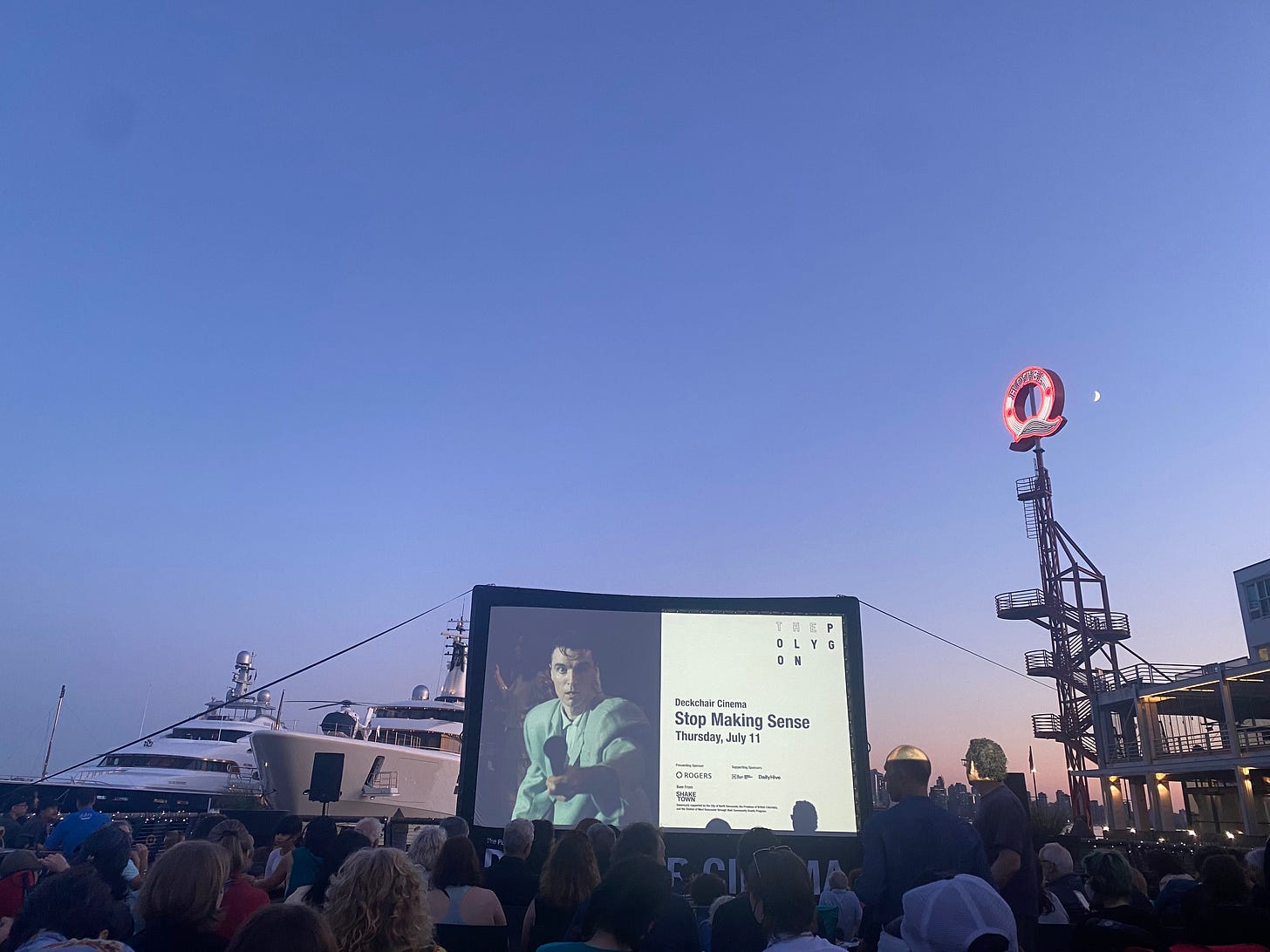 A blue hour photo of the screen at Deckchair Cinema, the 'Q' of Lonsdale Quay on the right, two large boats on the right. The moon is visible next to the lit-up Q. The screen is showing a promo for "Stop Making Sense" and the date Thursday, July 11. There are lots of heads visible in the foreground, people sitting in front of the screen.