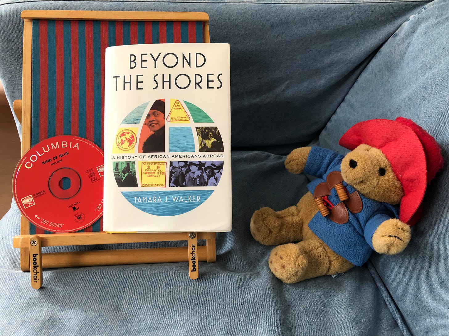 A CD (Miles Davis’s Kind of Blue) and a hardback book (Tamara J. Walker’s Beyond the Shores) sit on a bookchair next to a Paddington Bear teddy bear in a duffle coat and brimmed hat. The ensemble perches on the corner of a couch.