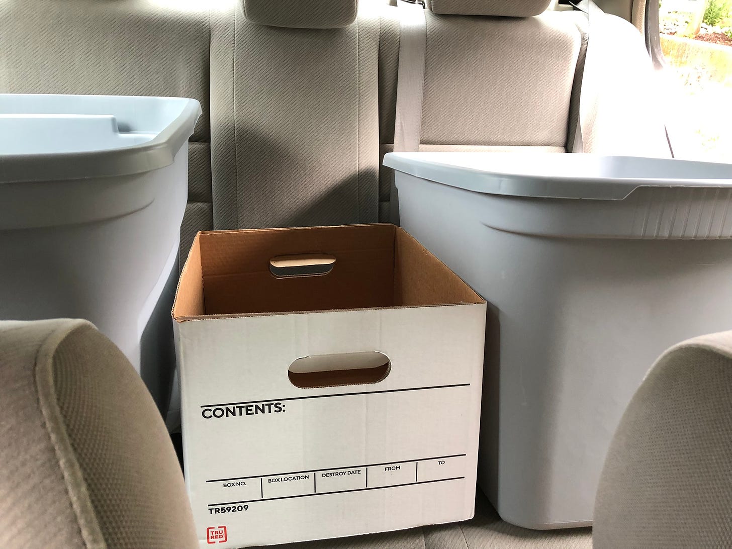 the beige backseat of a car filled with two large grey plastic bins and one staples brand carboard file box, no contents listed