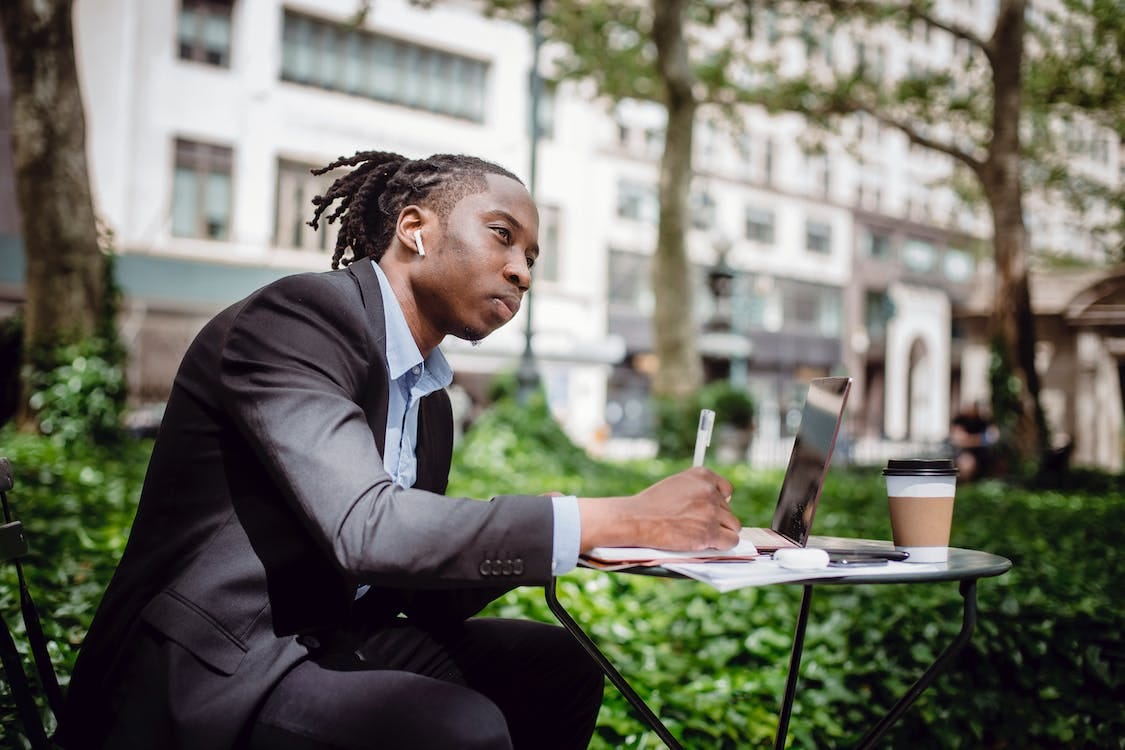 Free Side view of concentrated young black male writer listening to music with true wireless earbuds and looking away while creating new article in street cafe Stock Photo