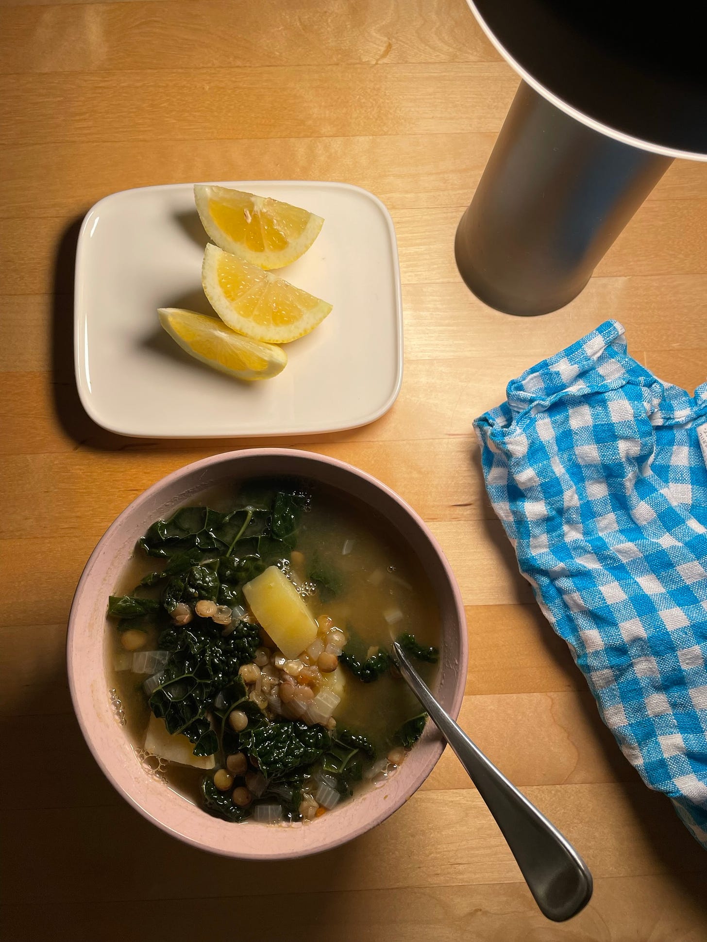 Pink bowl of lentil and potato soup with Tuscan kale. A plate of lemon wedges and a blue and white napkin sit nearby.