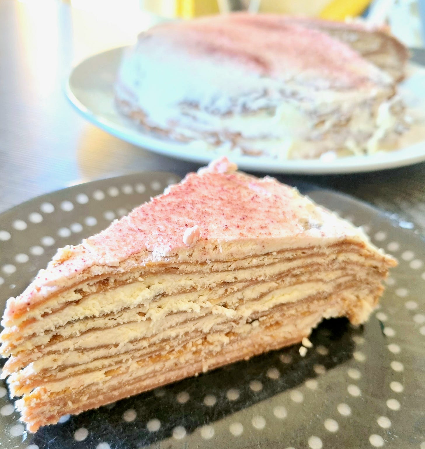 The image shows a slice of a layered cake on a plate with a dark background and white polka dots. The cake appears to have multiple thin layers, possibly indicating it is a crepe cake or a similar type of layered dessert. The top of the cake slice has a light dusting of what might be powdered sugar or another fine topping. In the background, there is another cake on a plate, which seems to be the whole cake from which the slice was taken. The background is slightly out of focus, emphasizing the slice in the foreground.