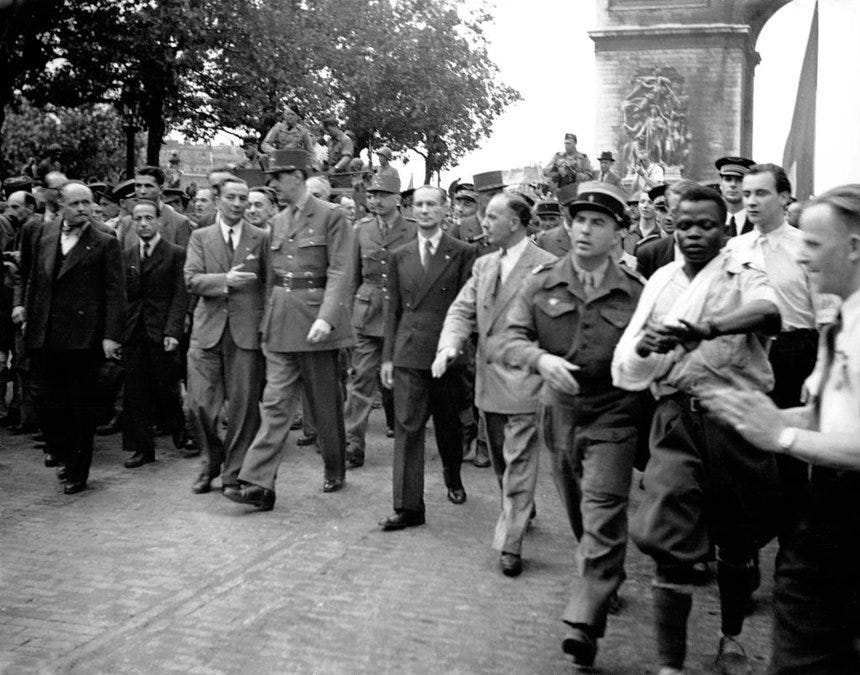 A black and white photo showing General Charles de Gaulle at the liberation of Paris parade. De Gaulle is surrounded by many other white men in suits and military uniform, but to the right of the frame there is a single Black man who appears to be getting pulled away but someone out of frame.