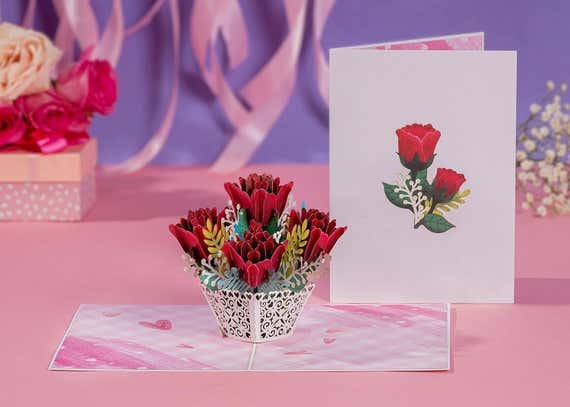 The Paper Love Lovely Roses Pop Up Card being displayed in a pink surface next to flowers and ribbons.