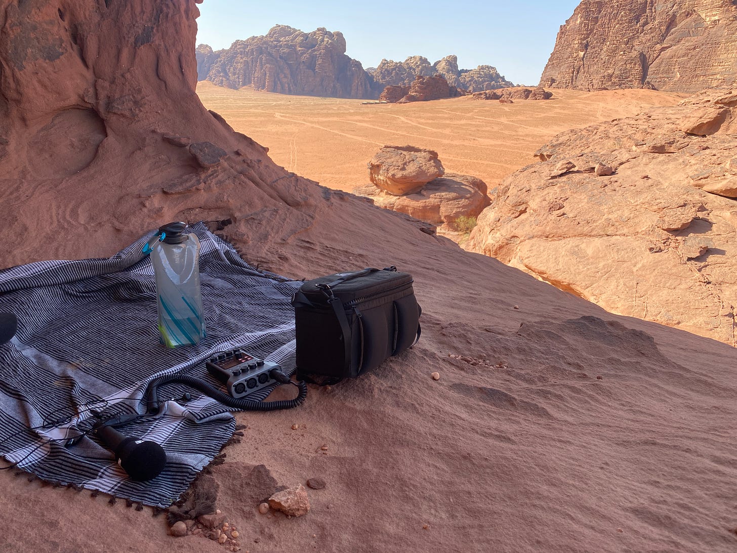 Our recording equipment in the Wadi Rum desert - a water bottle, a camera bag, a podcast recorded and a scarf as blanket. Desert rock vistas in the background.