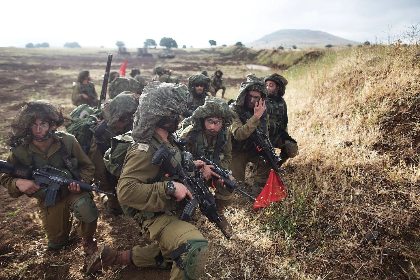 A group of about a dozen Israeli soldiers with an ultra-Orthodox Jewish battalion called Netzah Yehuda take part in a trainng operation on a cloudy but bright day. The soldiers are dressed in dark green camouflage and tactical face paint as they hold rifles and gesture to each other as they crouch to hide behind a grassy ridge. A muddy field stretches into the distance behind them.