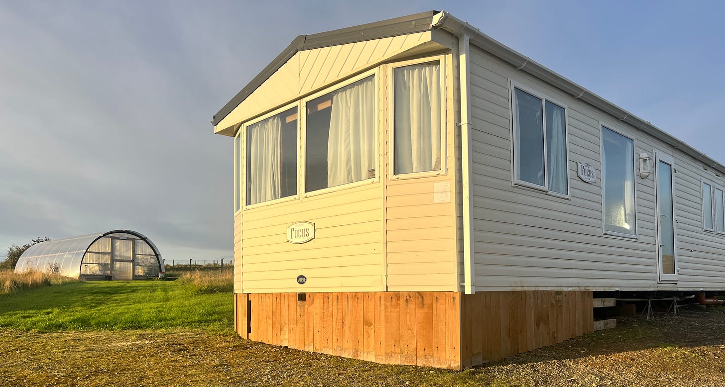 image of Colin the static caravan, with decking around the bottom. Doug the polycrub is in the background