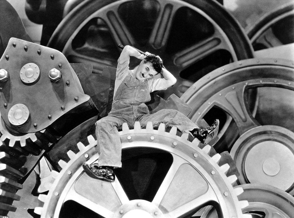 Imagem em formato quadrado, na cor preta e branca. No centro está Charles Chaplin sentado em cima de uma enorme engrenagem, com várias peças gigantes atrás. Ele está sorrindo com a mão atrás da cabeça. Ele é branco, usa um macacão de operário e sapatos grandes e desproporcionais..