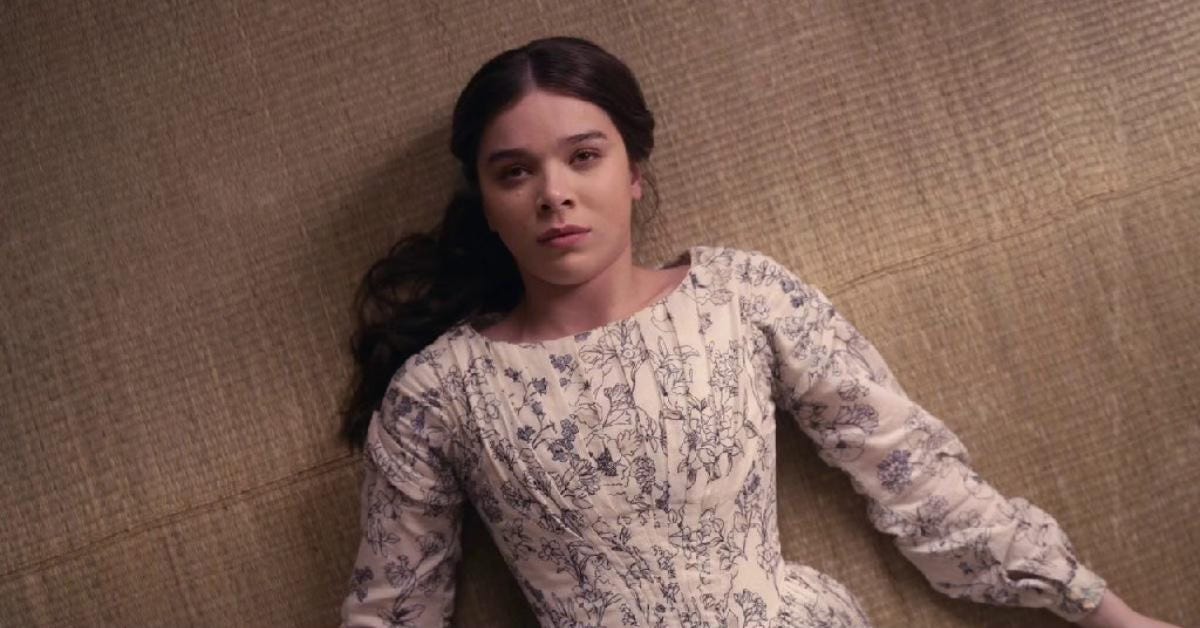 Still from the show Dickinson. Emily Dickinson lies on the floor in a black and white print dress, staring at a point above the camera.