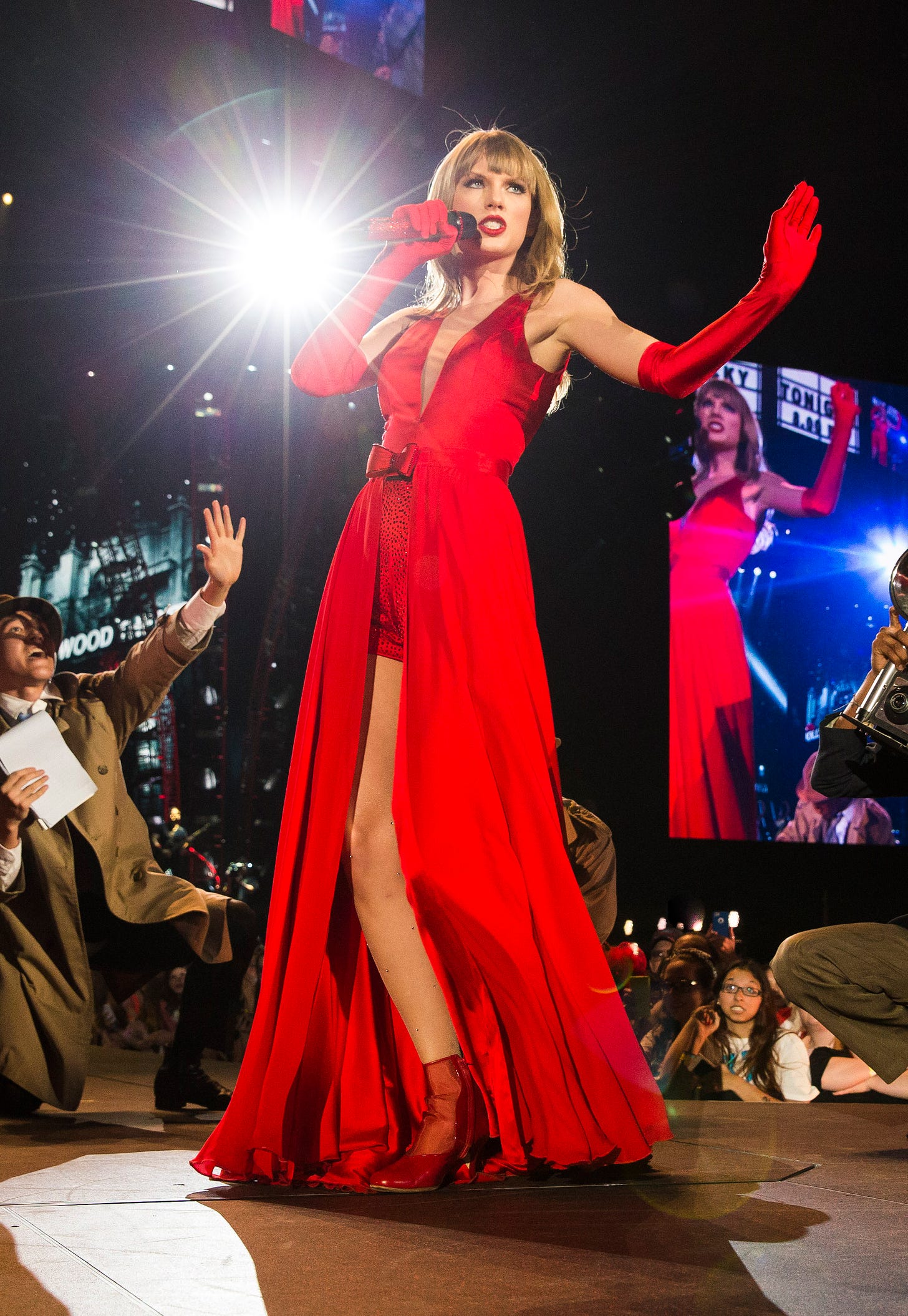 A young blonde woman with long hair in a floor length red dress holds a microphone