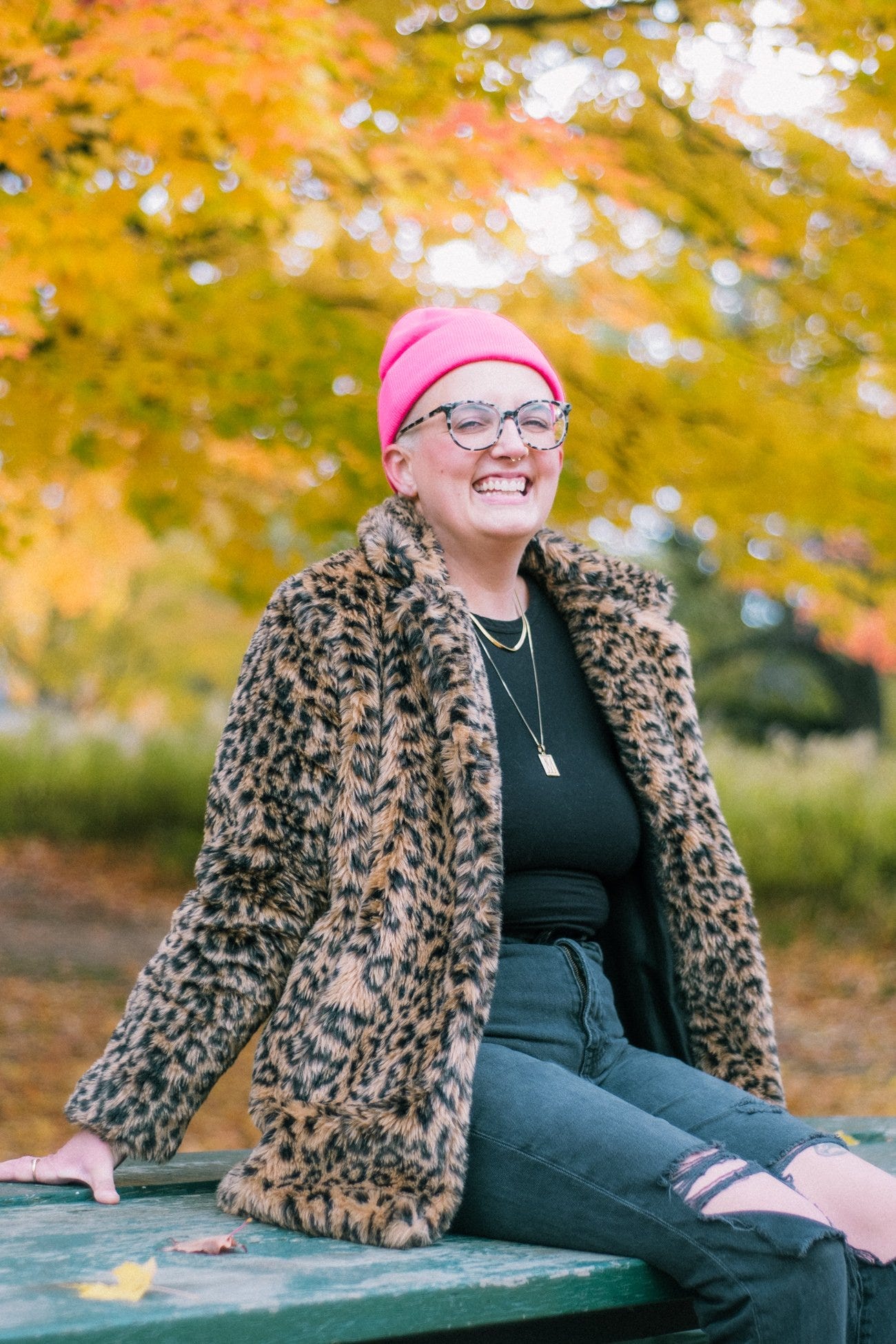 A professional portrait of Maia smiling wide outside in a park with orange and red autumn trees in the background, wearing glasses, a pink toque, and leopard print jacket.