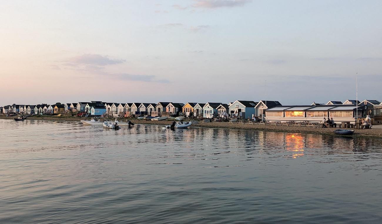 View of a row of beach huts with sun reflecting off them, with boats pulled up on the shore in front.