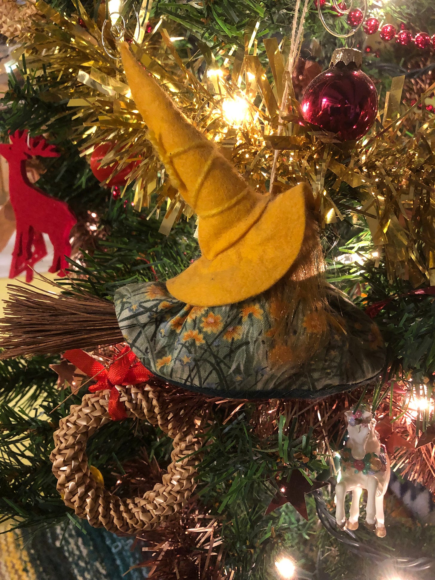 A little witch with a big yellow hat on a Yule tree. Other decorations are also visible: a red stag, some fairy lights, some tinsel, a Swedish straw ring, a shiny bauble, and a unicorn.