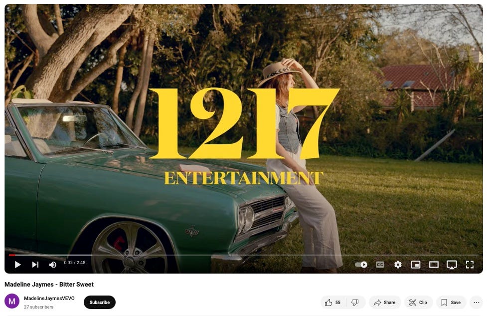 Screenshot of the "Bitter Sweet" music video on YouTube. Madeline is holding a wide-brimmed hat onto her head while posing on the hood of a motionless vintage car.