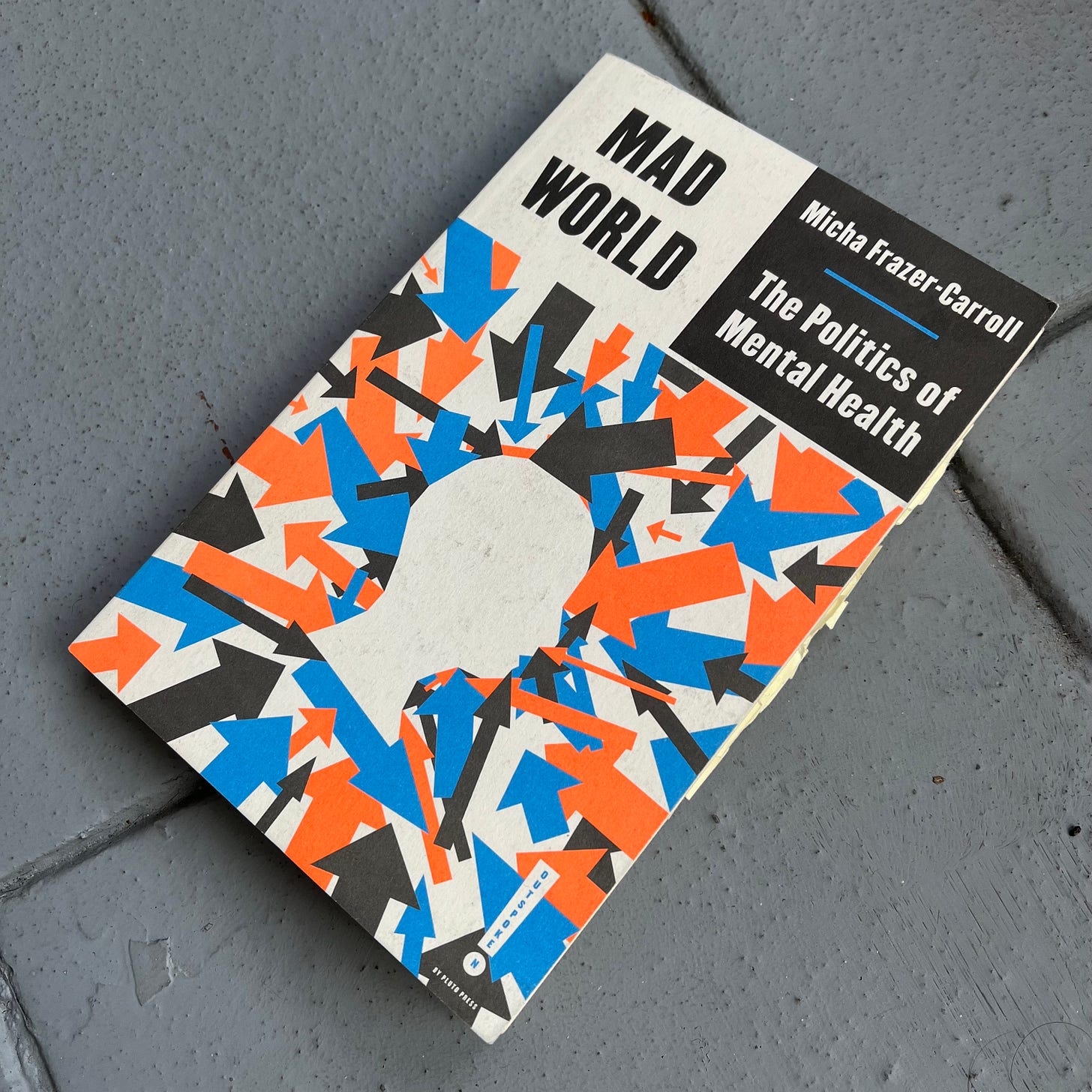 A picture of the book 'Mad World: The Politics of Mental Health' by Micha Frazer-Carroll. The book is resting on a grey tile floor.