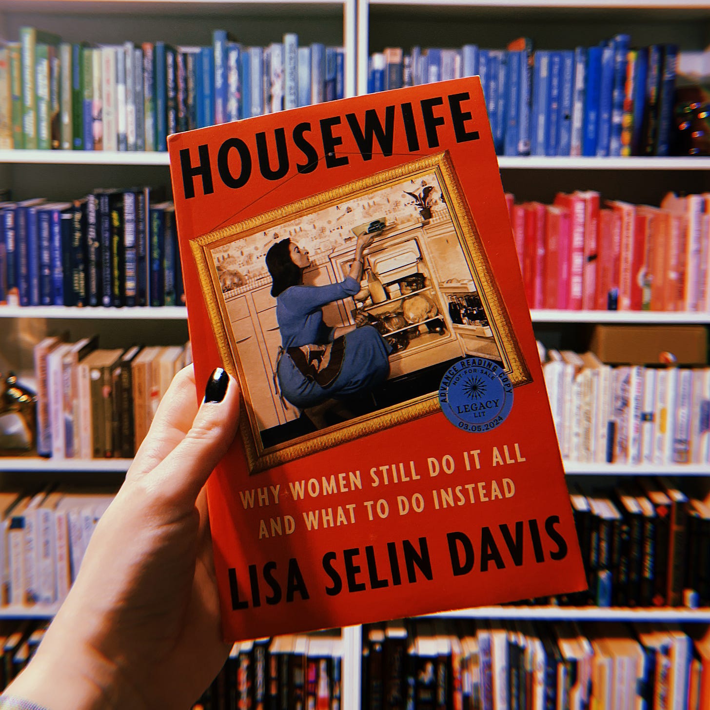 A hand holding up an advanced reader copy of Housewife: Why Women Still Do It All and What to Do Instead by Lisa Selin Davis. A bookshelf is in the background.