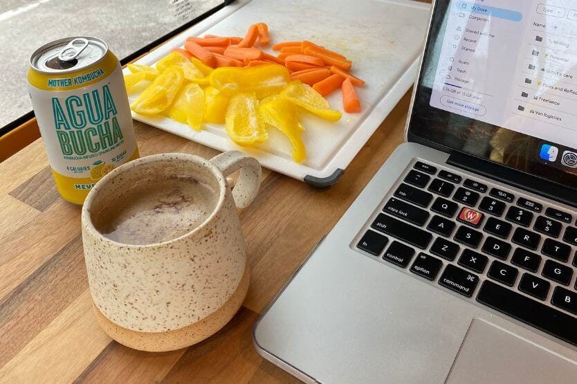 A mug of coffee, a can of kombucha, and some sliced vegetables sit on the butcher block of our converted camper van next to my laptop