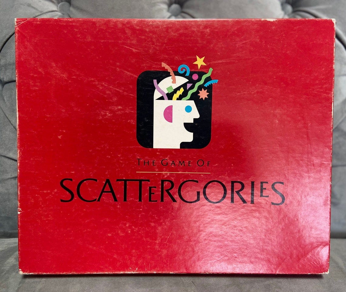 May include: The game of Scattergories box is red with a white head with a blue eye and a red ear. The head is surrounded by confetti and stars. The text on the box reads 'The Game of Scattergories'.