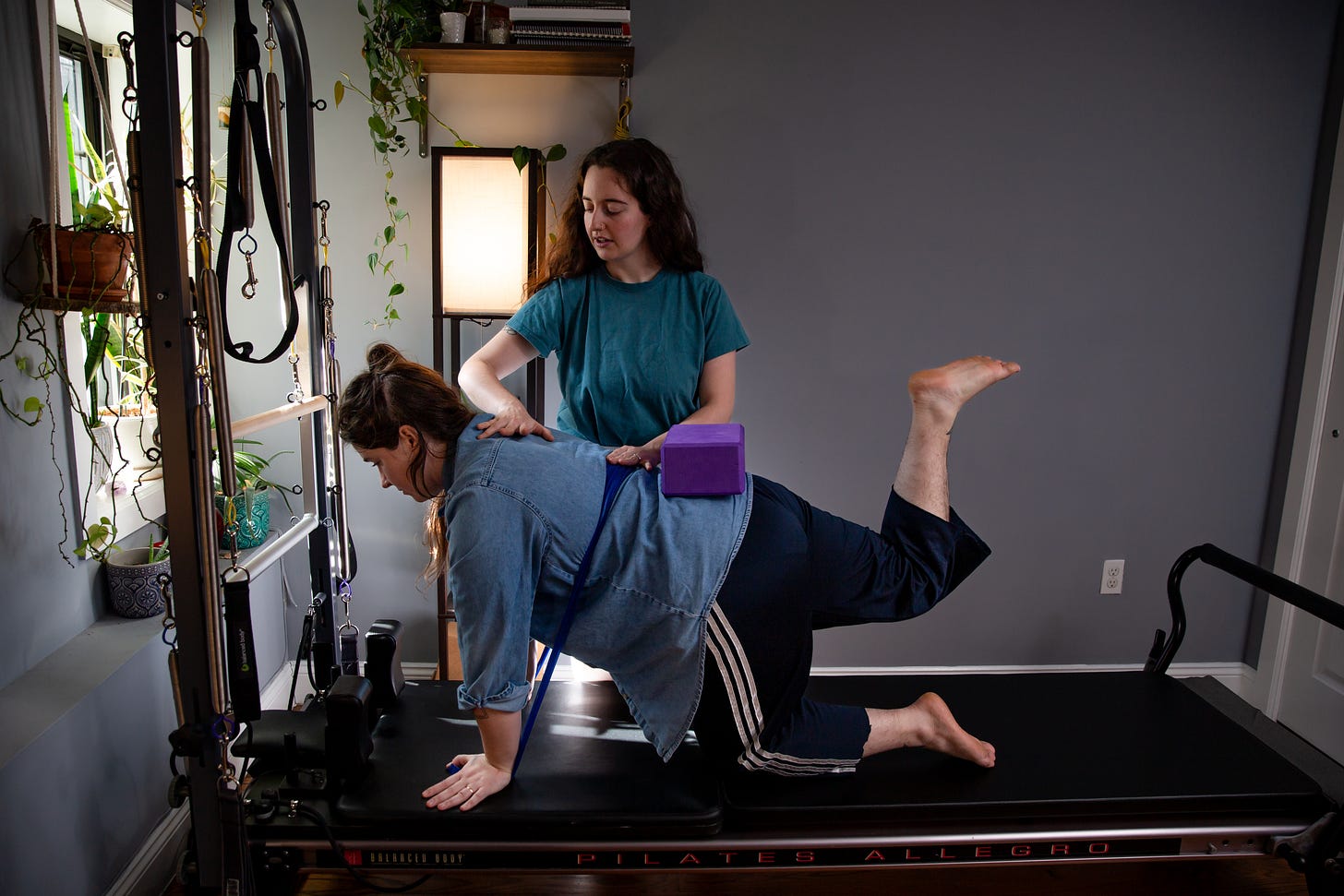 Sofia, who has long brown hair and wears a blue t-shirt, guides a client through movement on the Pilates reformer, placing her hands gently on the client's back.