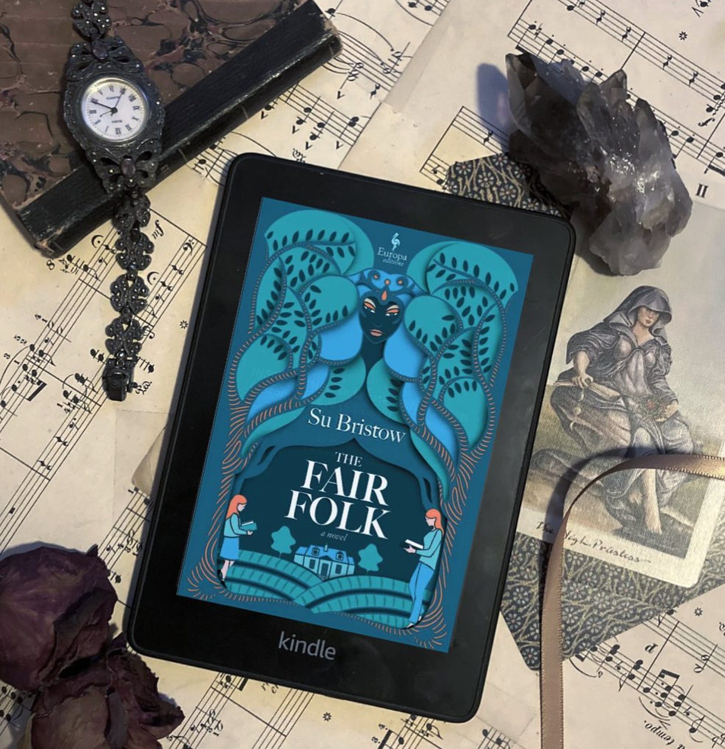A flat lay of The Fair Folk on a Kindle screen with a background of sheet music, a 1940s-style marcasite Art Deco watch, dried roses, crystals, tarot cards and a ribbon.