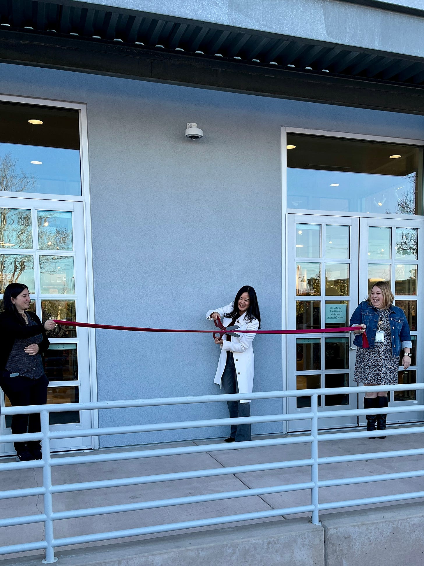 Author Evelyn Skye in white coat, holding giant scissors and cutting a red ribbon to open a Barnes & Noble bookstore