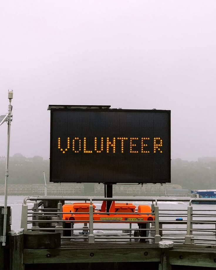 a black construction sign that says VOLUNTEER in orange lettering against a gray and foggy worksite backdrop