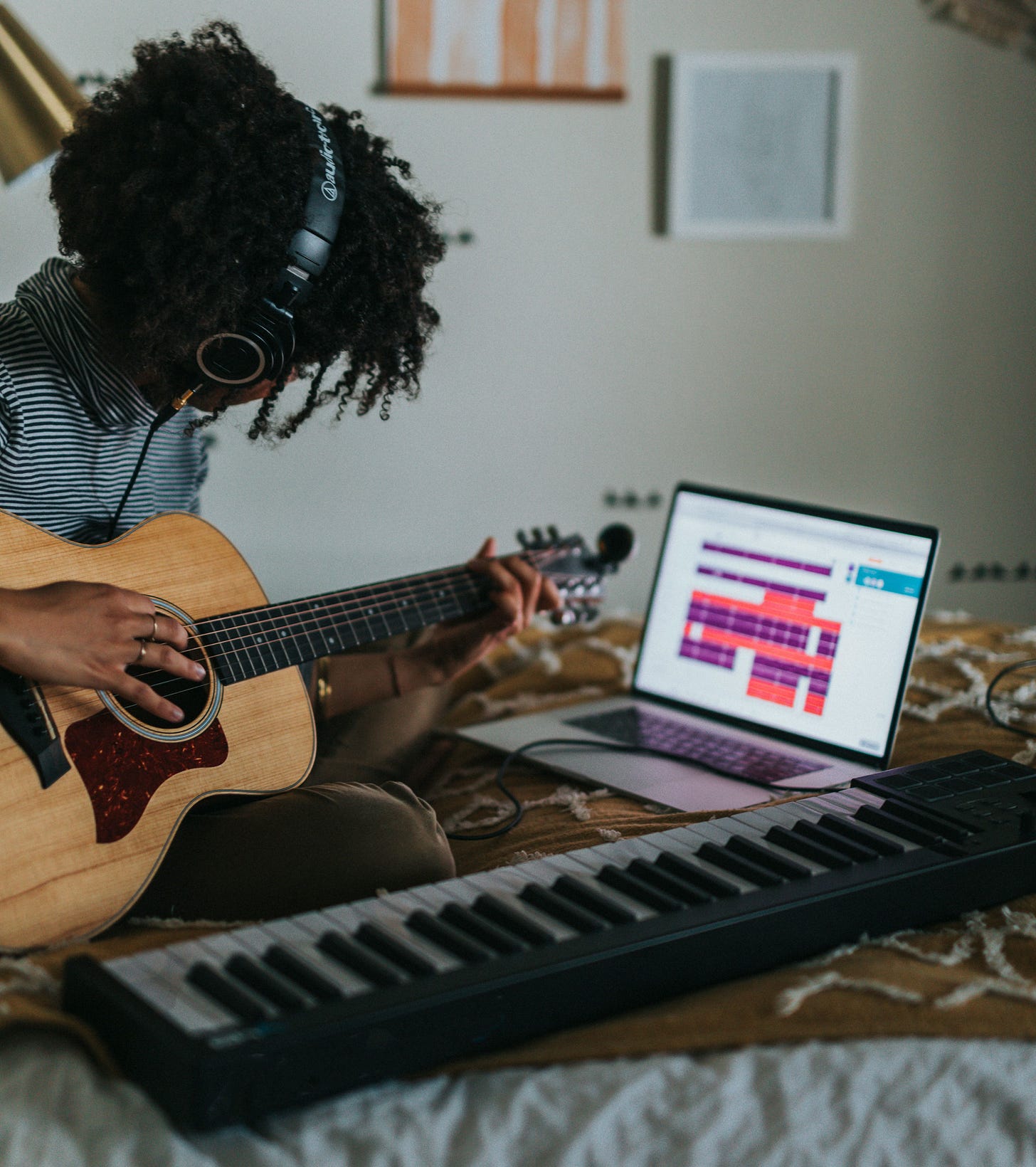 Black woman sitting on a bed wearing headphones, playing a guitar, looking at music on a computer, with a music keyboard nearby