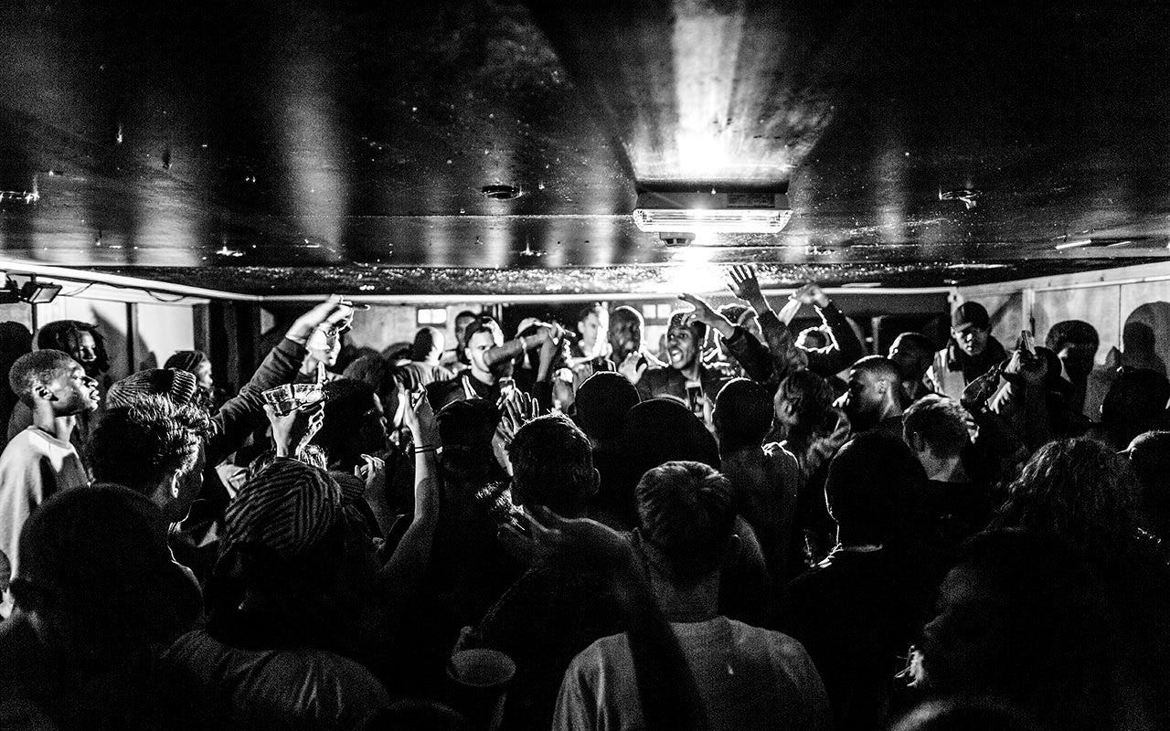 Black and white photo in a basement nightclub full of young people partying with their hand in the air.