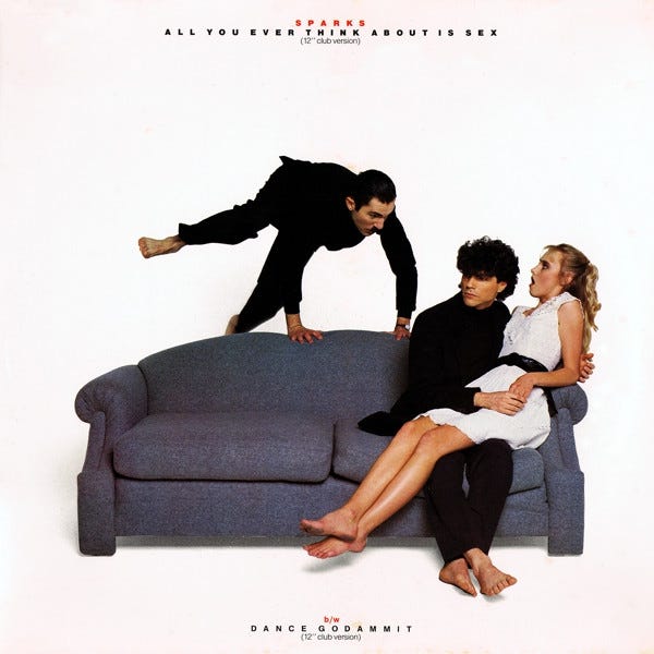 Ron Mael leaps over a grey sofa towards Russell Mael who has a woman in a short white dress on his lap.