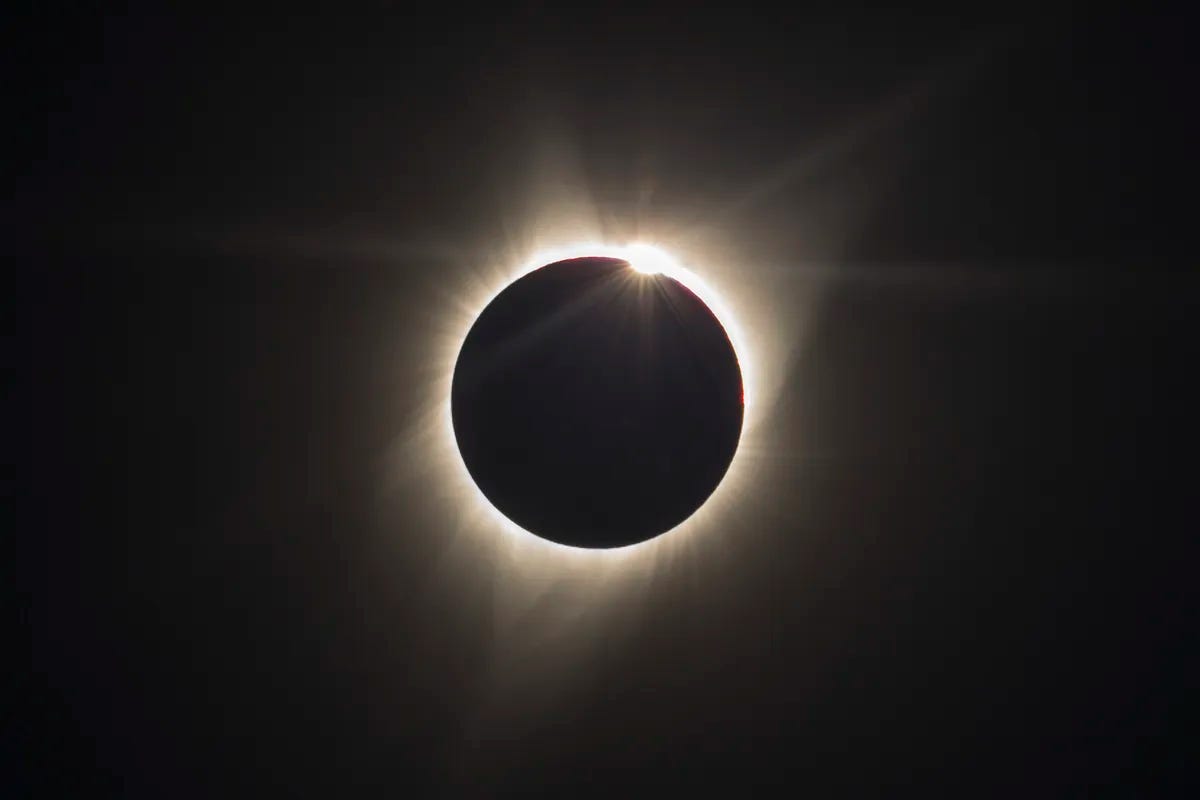 A solar eclipsed is shown with the black disk of the moon completely eclipsing the Sun. A white halo of light surrounds the black disk of the moon and a dot of light--known as a Bailey's Bead--shines on the upper right part of the sun. 