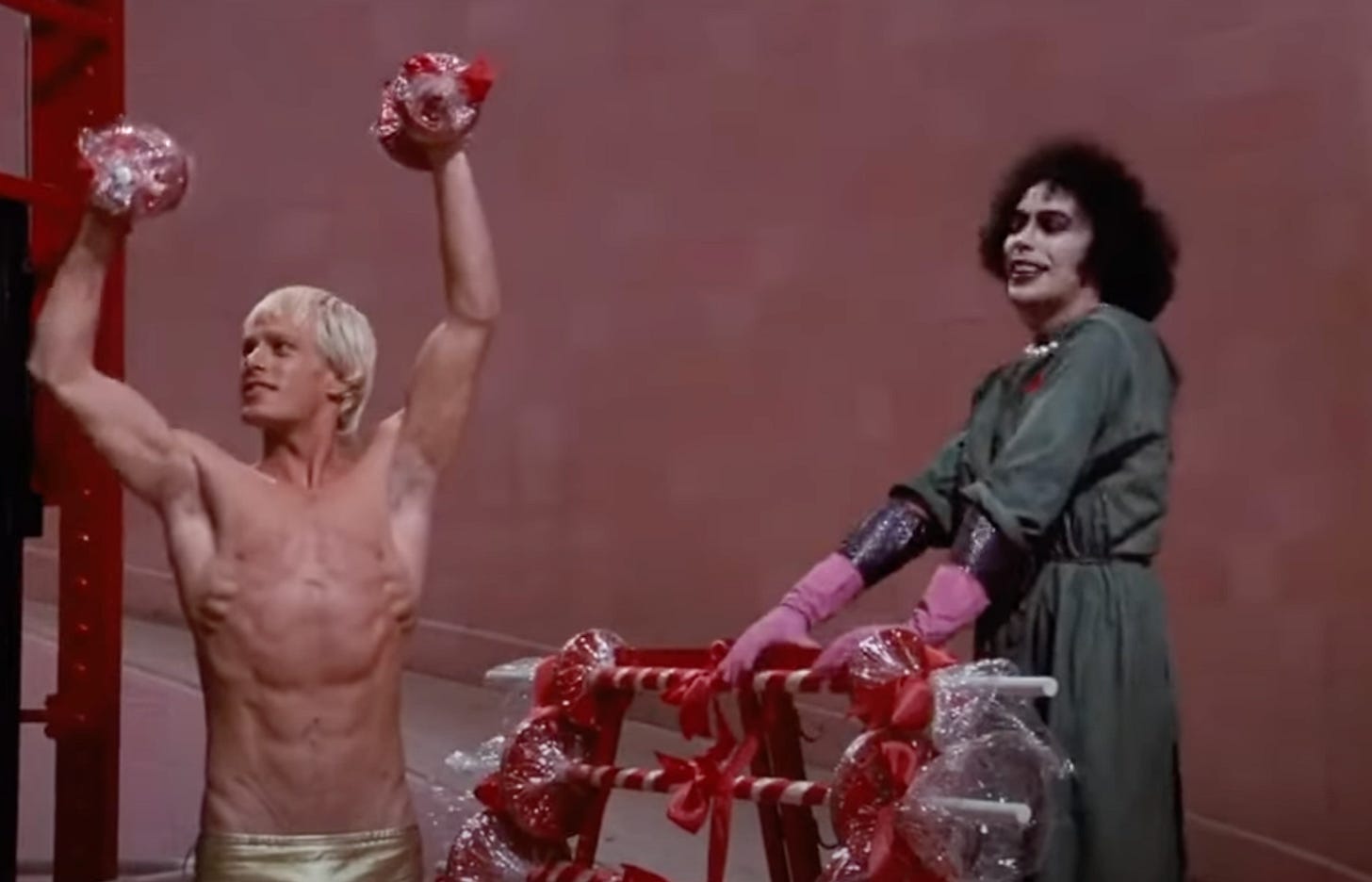 Still from Rocky Horror, from the I Can Make You a Man scene. Rocky holds up weights in the air and shows off his muscles while Tim Curry stares at him lasciviously