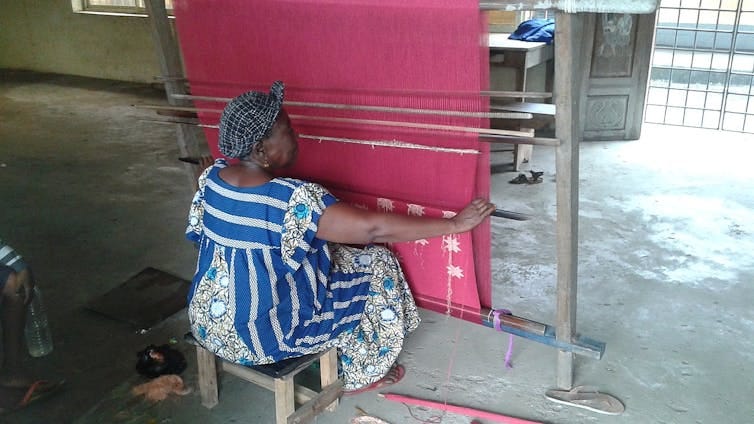 A woman in a bright blue dress in traditional fabrics sits at a weave working on a vast drop of bright pink threads. A woman in a bright blue dress in traditional fabrics sits at a weave working on a vast drop of bright pink threads.