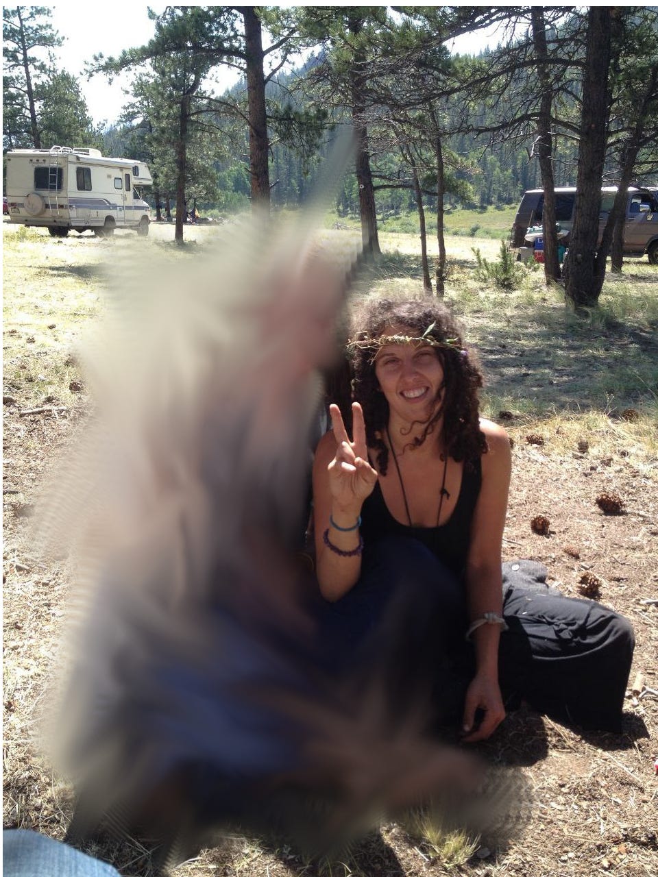A photo of the author wearing a Crown of vines in a Colorado forest clearing, making a V sign with one hand, sitting cross-legged on the ground next to another person who is blurred out for anonymity purposes.