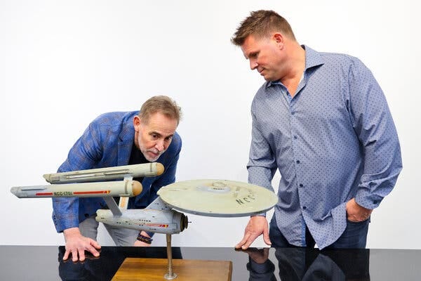 Two men, both wearing blue shirts, inspect a model of the U.S.S. Enterprise.