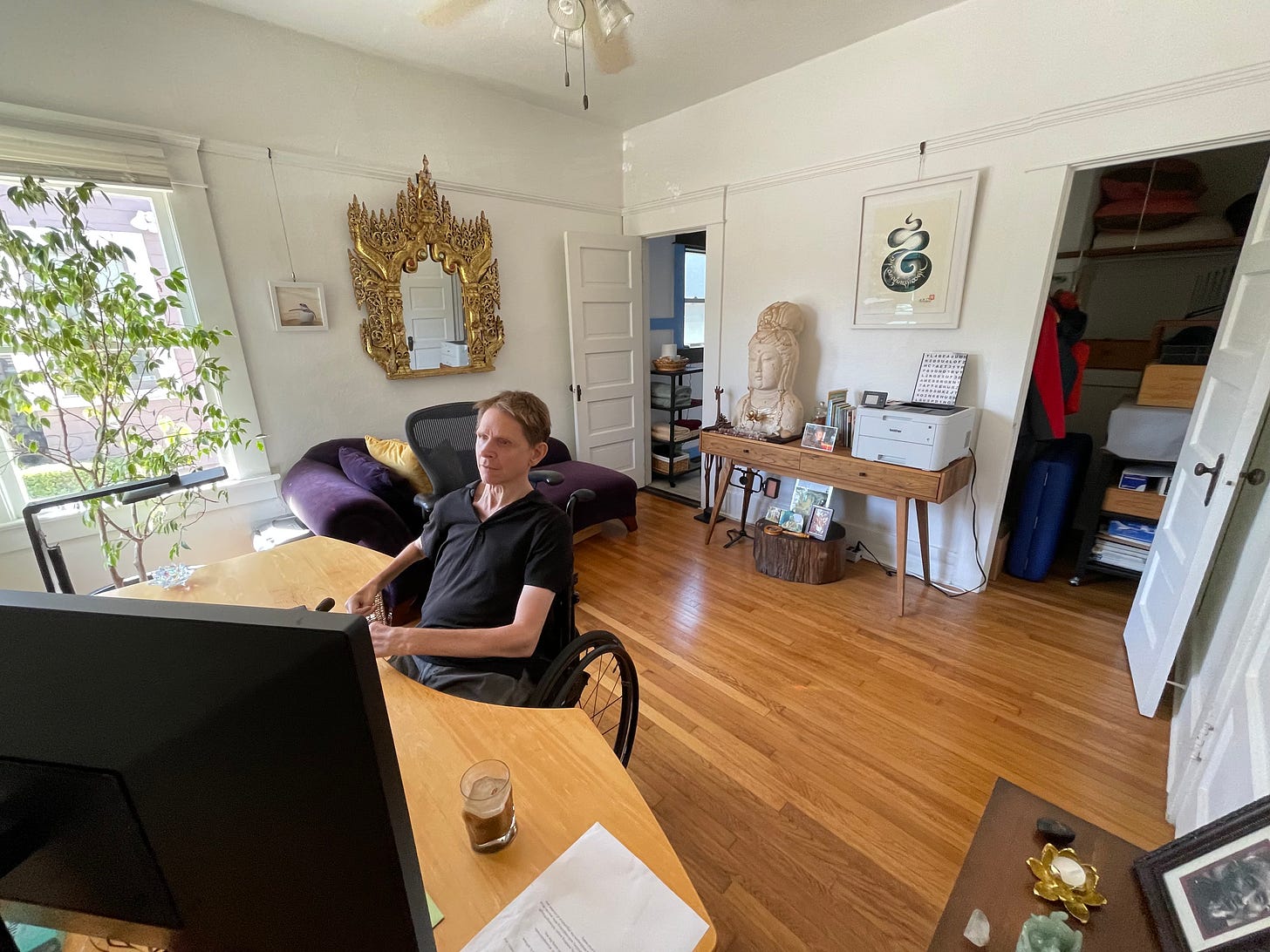 Kabir at home at work in his office. A small light filled room, a small tree in the corner to the left, a gold and ornate mirror on the wall, and Buddhist sculpture and printer on a small table along the back wall. To the right frame, to the left of Kabir as he sits at his desk, a small altar with a photo of Geshe Wangyal.