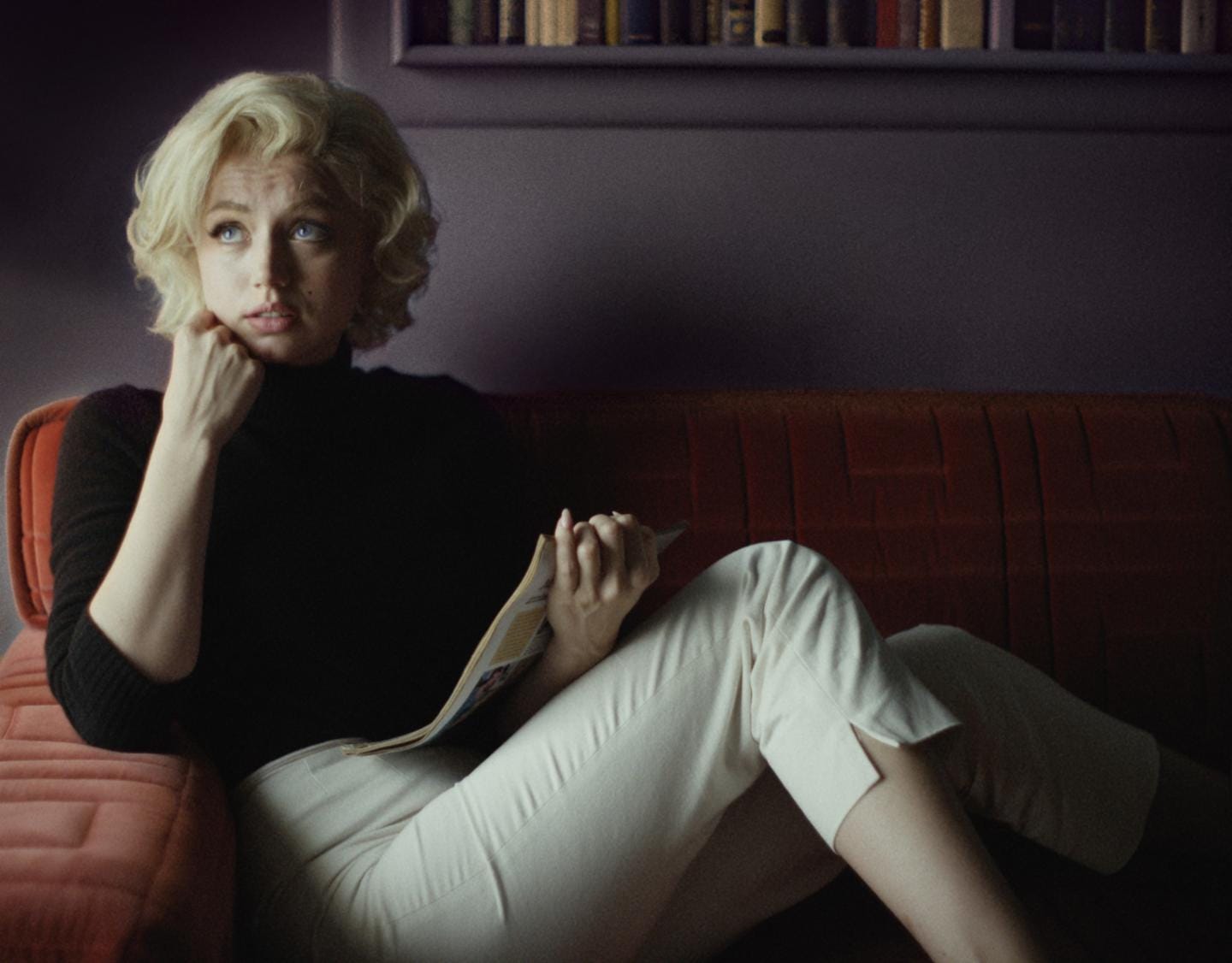 A white woman with blond hair sits on a sofa. She is wearing a black turtleneck and white pants.