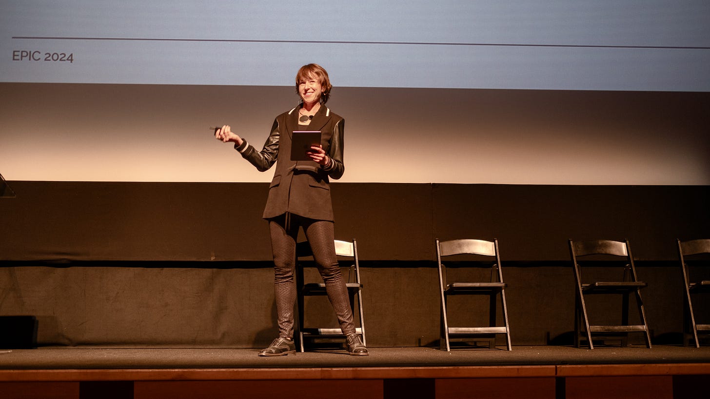 Tall woman with medium-length brown hair stands on a stage. She holds a clicker in one hand and an iPad in the other. She is dressed in a black sport coat blazer, black tights with a pattern and black shoes.